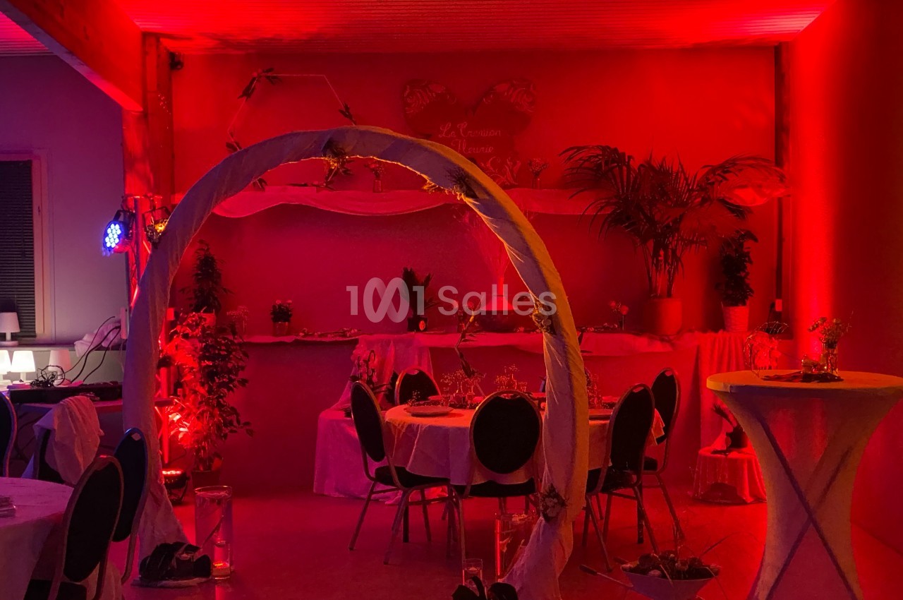 Salle décorée pour un événement, éclairée par une lumière rouge avec tables rondes, plantes et une arche ornée.