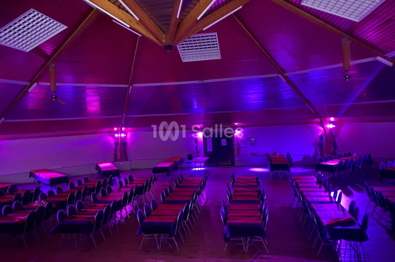 Salle de réception avec tables alignées, éclairage tamisé violet et plafond en bois avec poutres apparentes.
