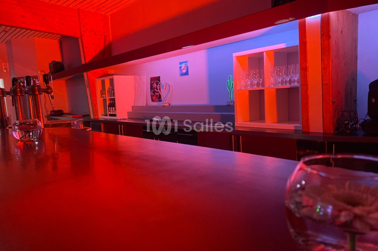 Bar avec éclairage rouge, comptoir en bois, étagères avec verres et décorations, ambiance tamisée.