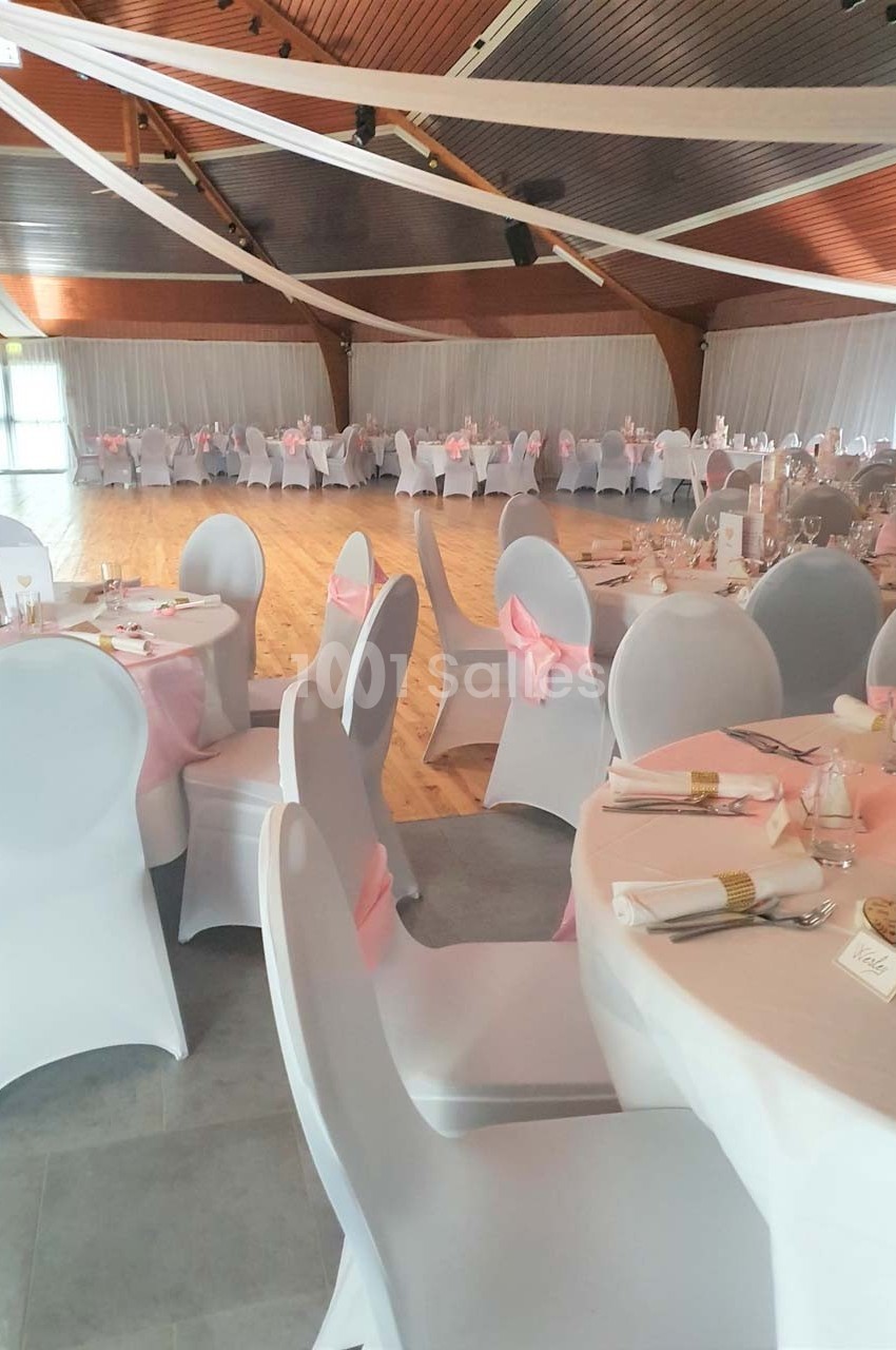 Salle de réception décorée avec des tables rondes, nappes blanches, chaises houssées et rubans roses.