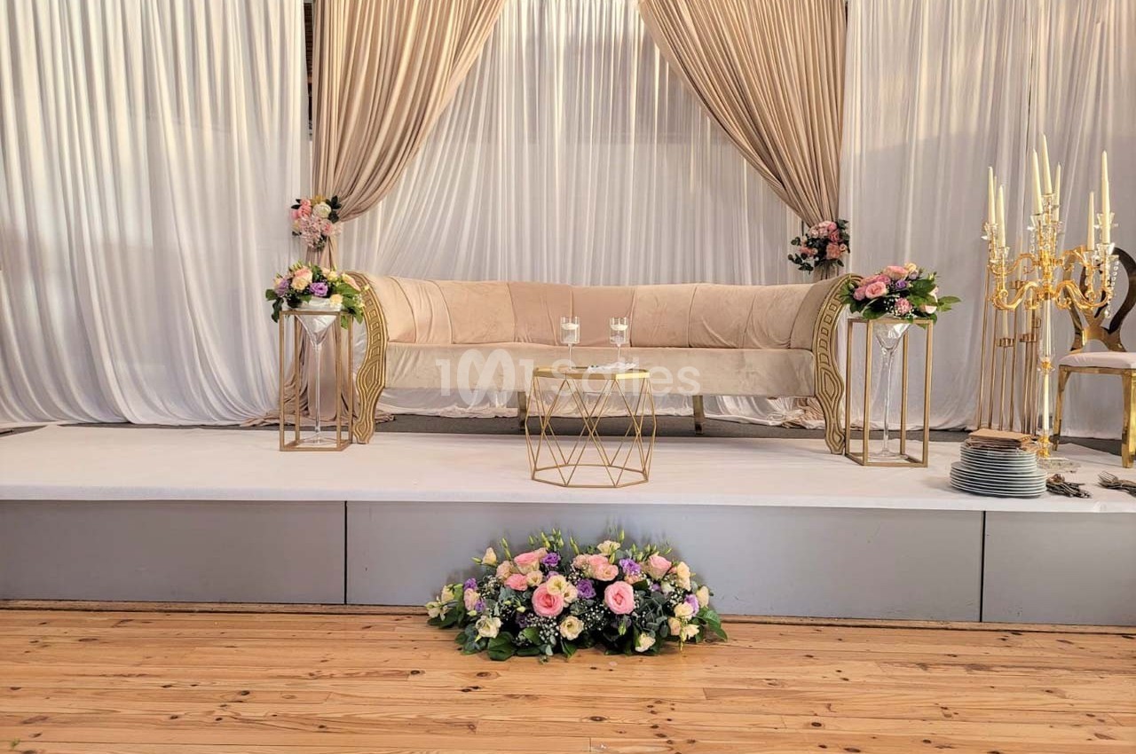 Canapé beige décoré de fleurs pastel, entouré de rideaux dorés et blancs, sur une estrade en bois.