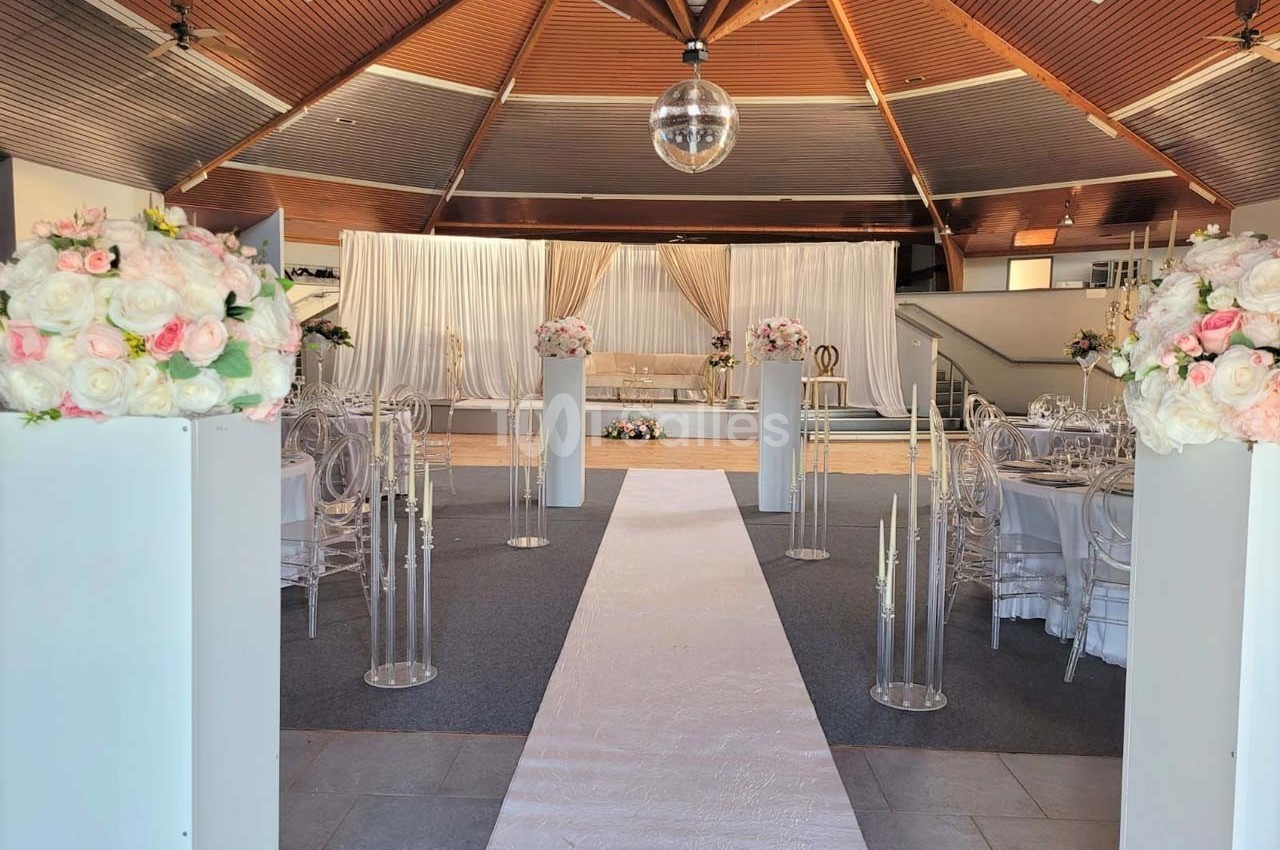 Allée centrale décorée de fleurs et chandeliers, menant à une scène avec rideaux et chaises dans une salle de réception.