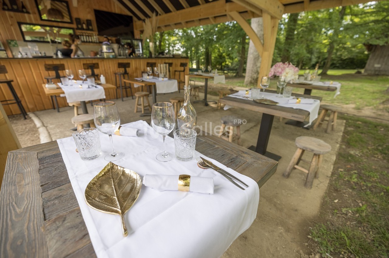 Tables dressées avec nappes blanches, couverts dorés et verres, dans un restaurant en plein air entouré de verdure.