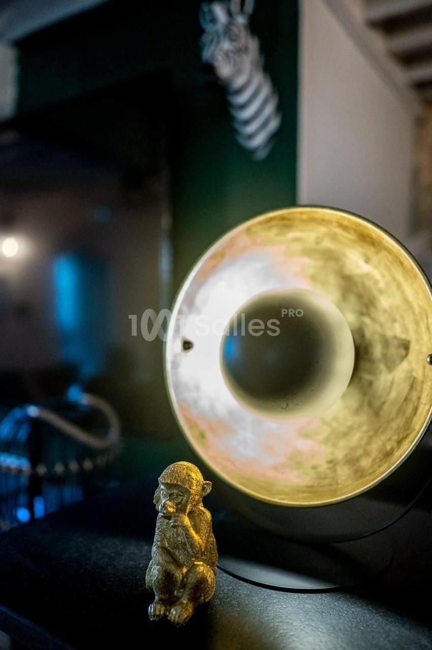 Lampe ronde illuminée avec une statuette dorée de singe au premier plan, décor intérieur sombre en arrière-plan.