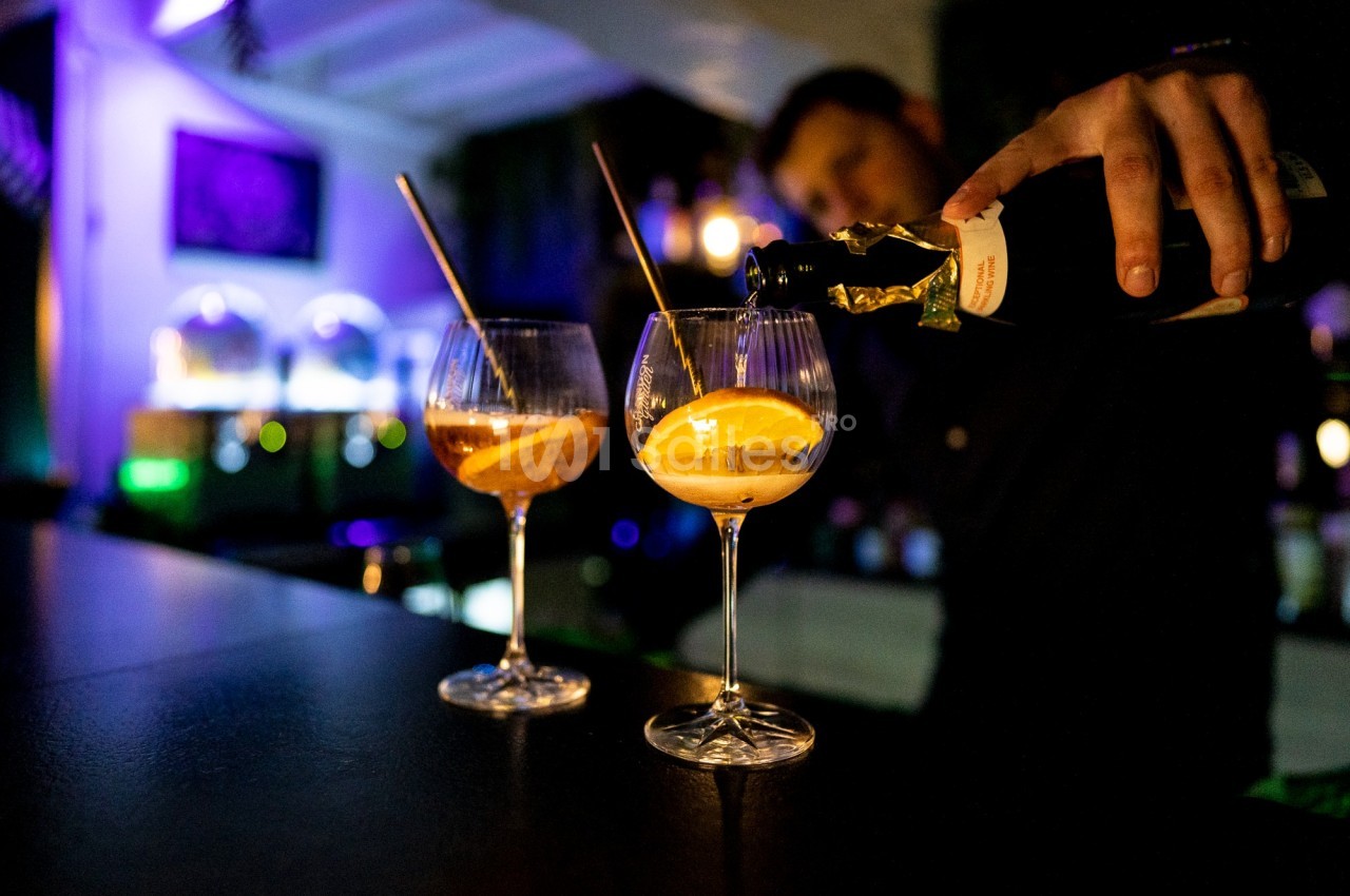 Un barman verse du champagne dans deux verres à cocktail garnis de tranches d'orange, dans un bar éclairé.