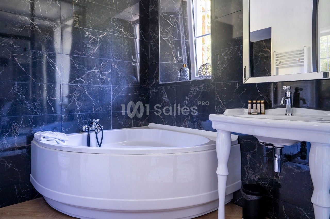 Salle de bain avec baignoire d'angle blanche, murs en marbre noir, lavabo sur pied et fenêtre laissant entrer la lumière.