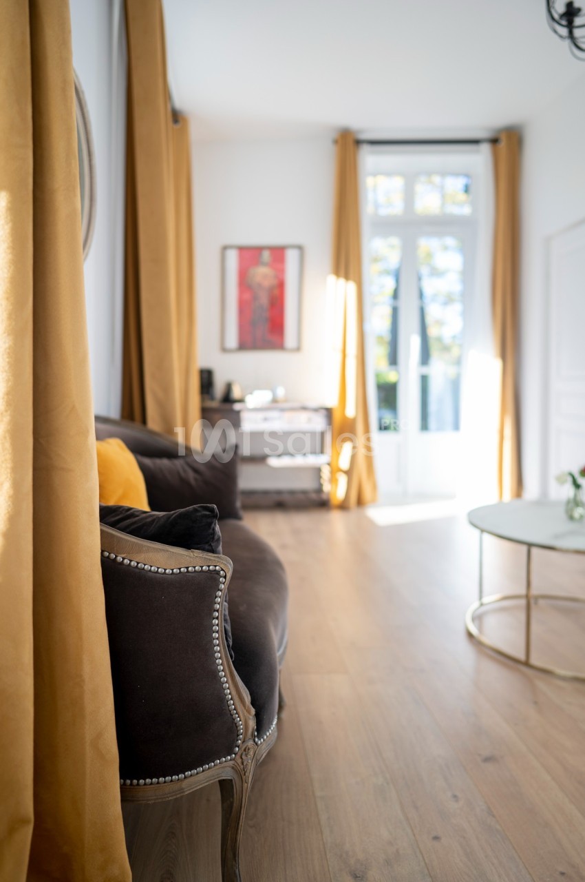 Salon lumineux avec canapé sombre, rideaux jaunes, table basse ronde et grande fenêtre donnant sur l'extérieur.