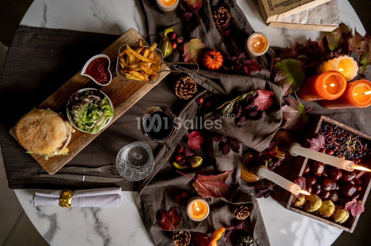 Table décorée avec des bougies, feuilles d'automne et un repas comprenant un burger, des frites et des accompagnements.