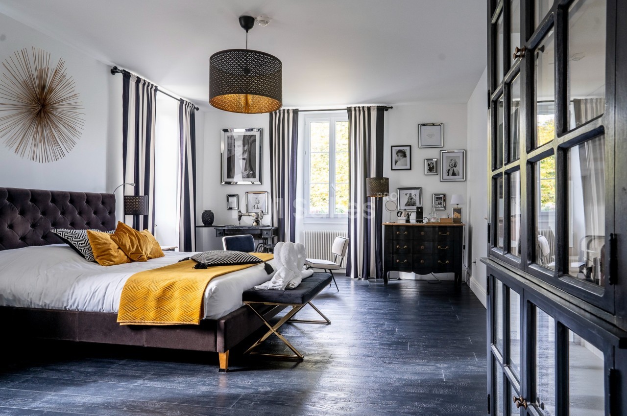 Chambre lumineuse avec lit double, décoration moderne, parquet sombre, murs blancs et grande fenêtre avec rideaux.