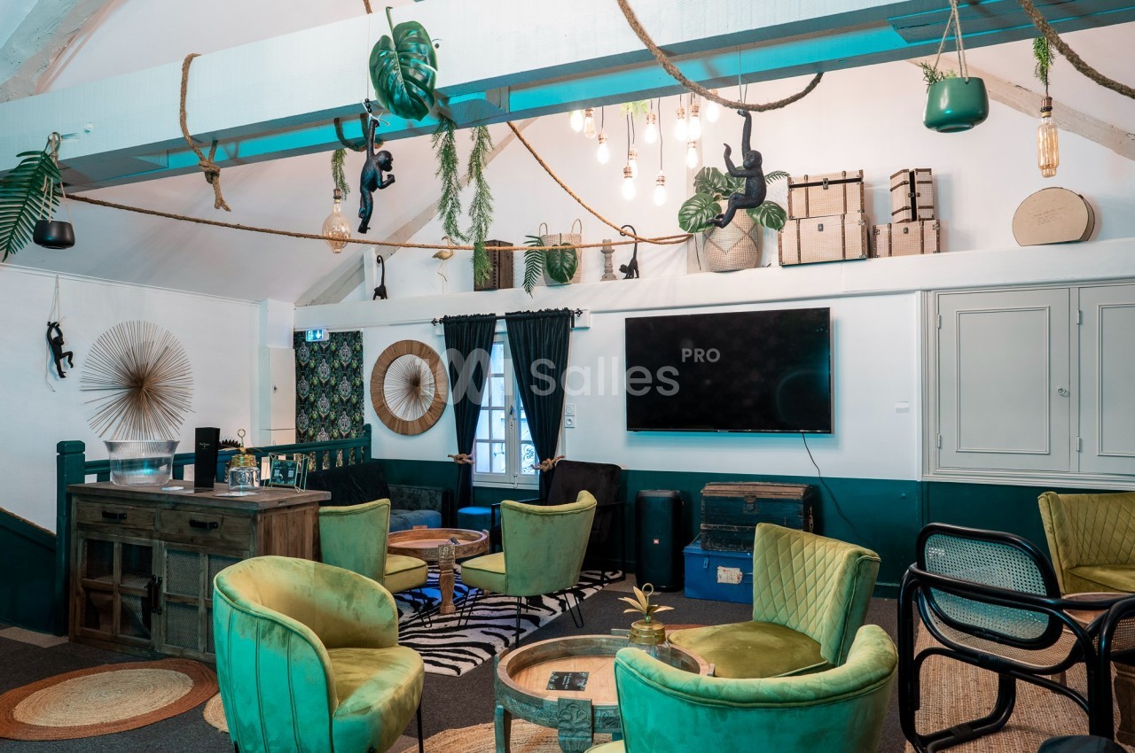 Salle de séjour lumineuse avec fauteuils verts, décor tropical, télé murale et éléments suspendus au plafond.
