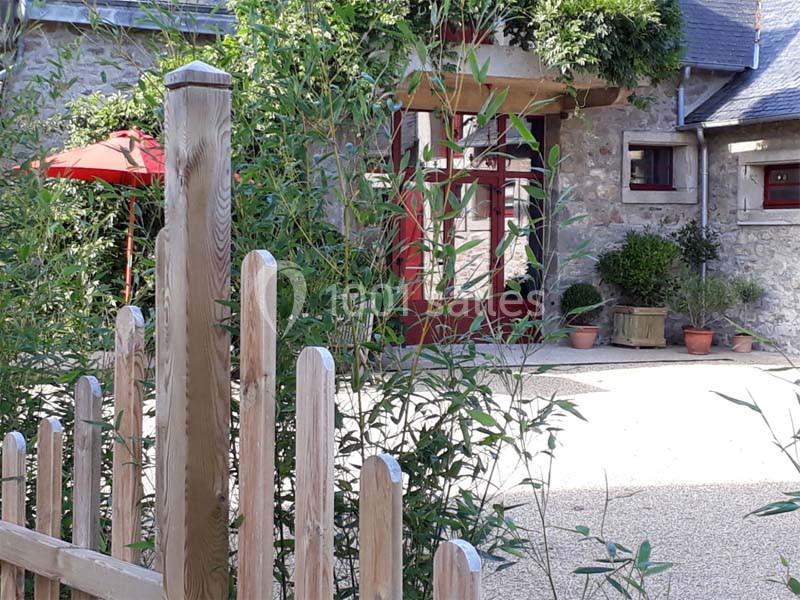 Façade en pierre d'une maison avec des volets rouges, un jardin arboré et une clôture en bois au premier plan.