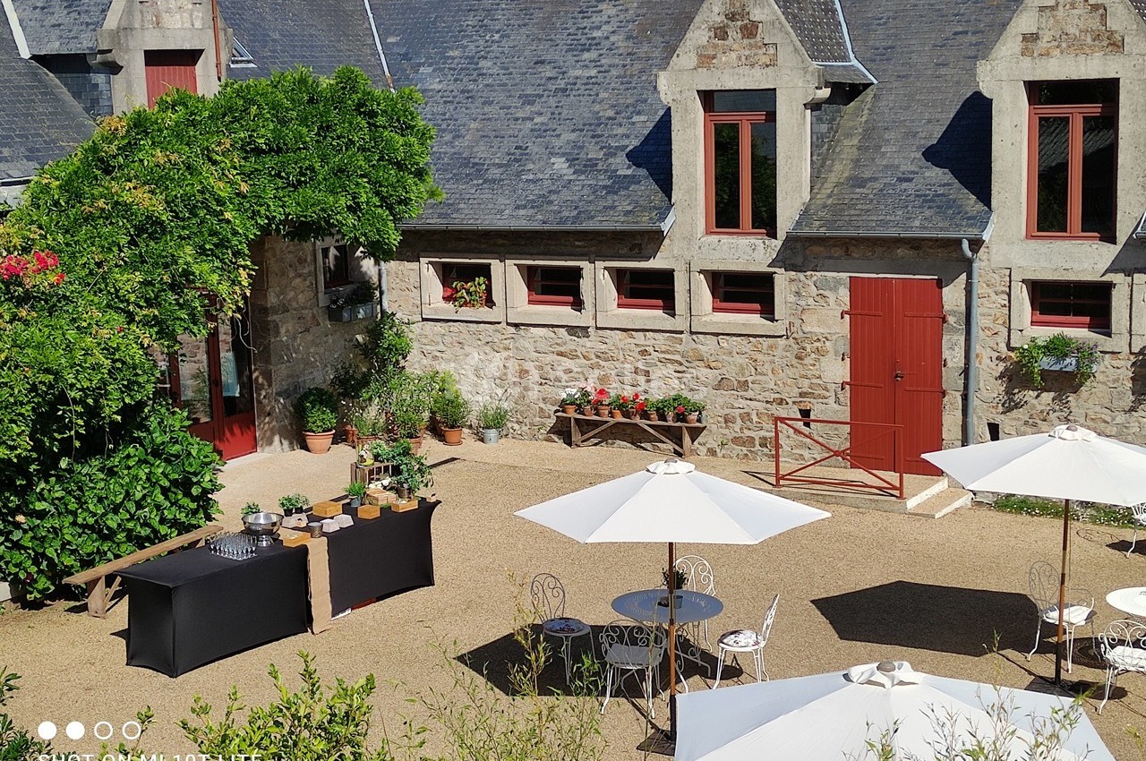 Cour extérieure ensoleillée avec tables, parasols blancs, plantes et bâtiment en pierre aux volets rouges.