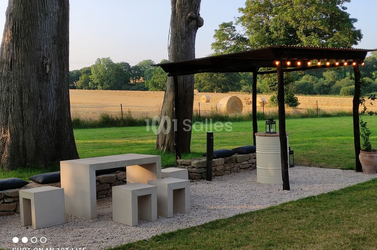 Espace extérieur avec table et bancs modernes sous une pergola éclairée, entouré de pelouse et de champs.