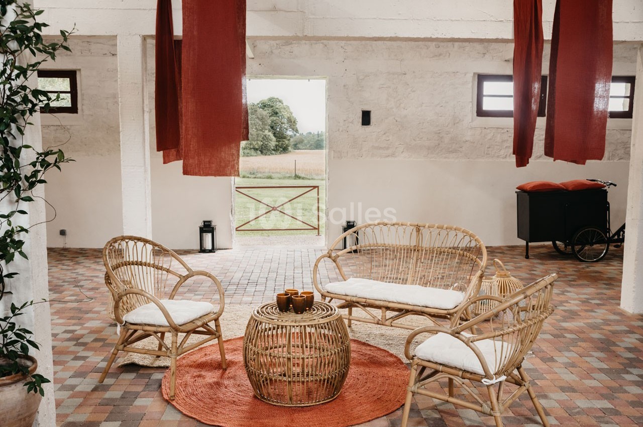 Espace intérieur lumineux avec mobilier en rotin, tapis orange et vue sur un jardin à travers une porte ouverte.