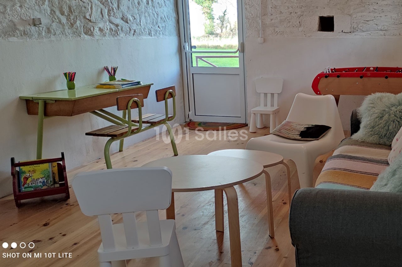Salle lumineuse avec mobilier simple : table, chaises, canapé, baby-foot et vue sur un jardin par une porte vitrée.