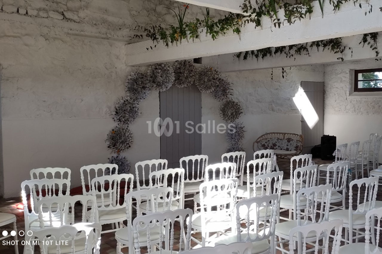 Salle décorée pour un événement, avec des chaises blanches alignées et une arche florale devant une porte grise.