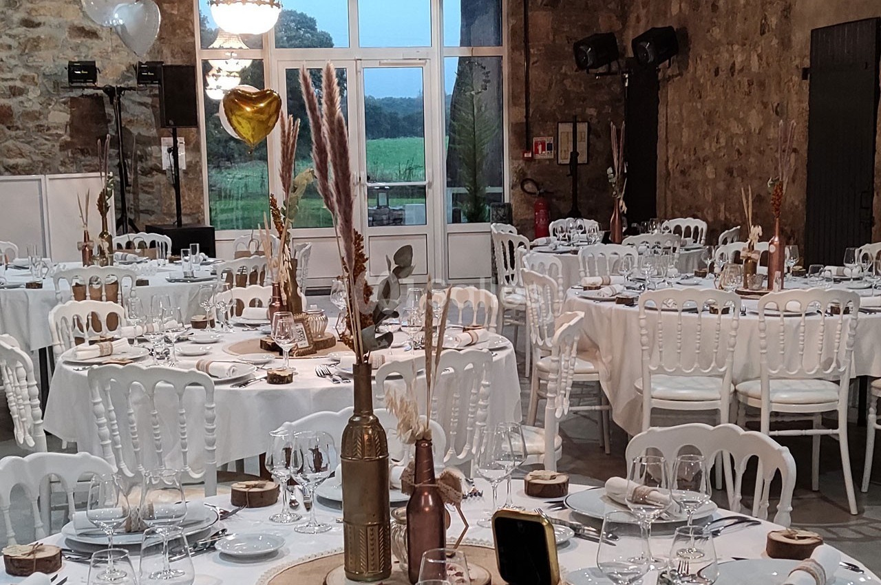 Salle de réception décorée avec des tables rondes, nappes blanches, chaises assorties et ornements champêtres.