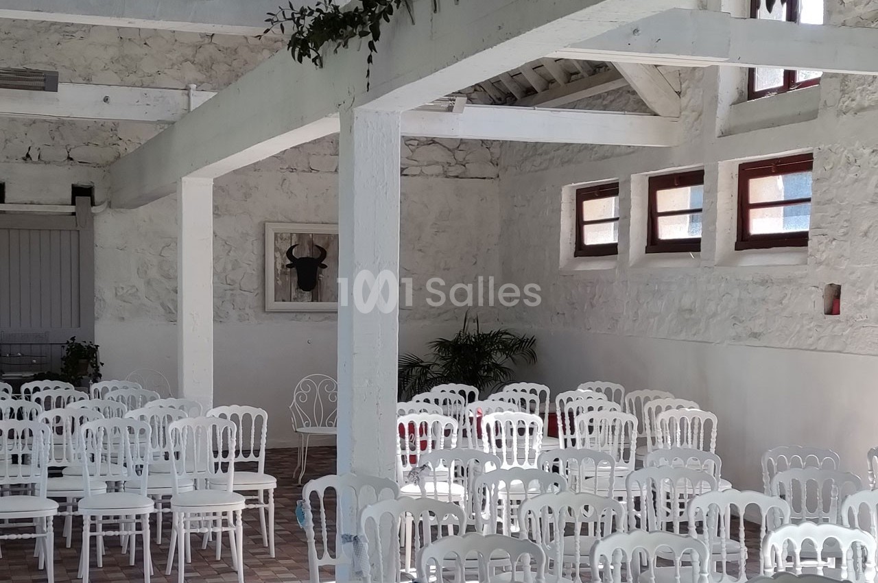 Salle lumineuse avec des murs en pierre blanche, des chaises blanches alignées et des poutres apparentes.