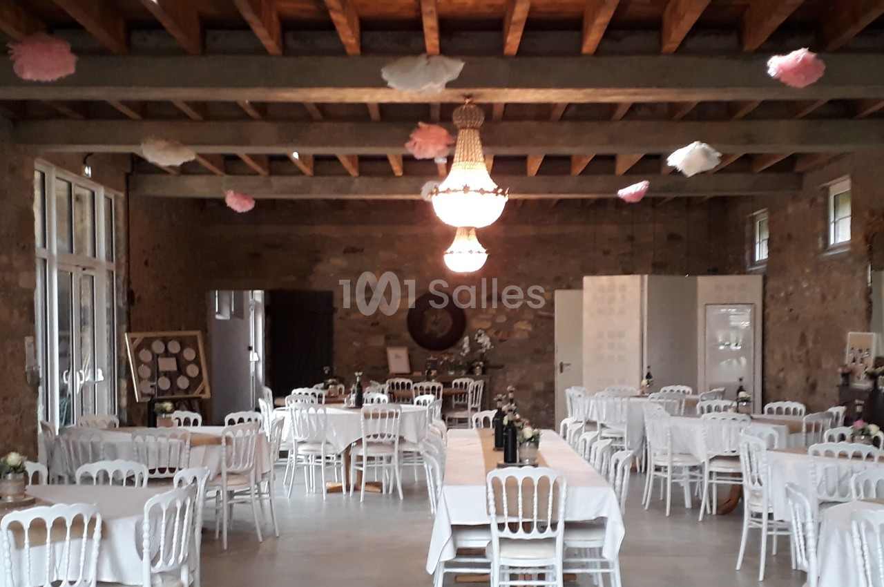 Salle de réception avec tables dressées, chaises blanches et décoration suspendue sous un plafond en poutres apparentes.