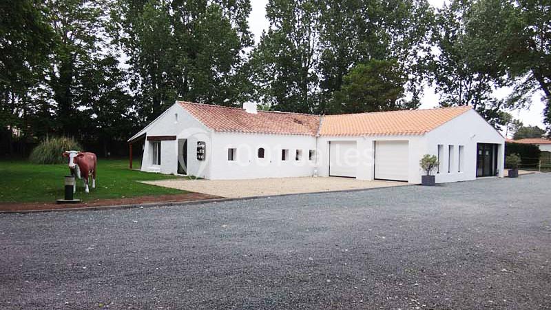 Maison blanche avec toit en tuiles orange, garage double, allée en gravier et statue de vache à gauche, entourée d'arbres.