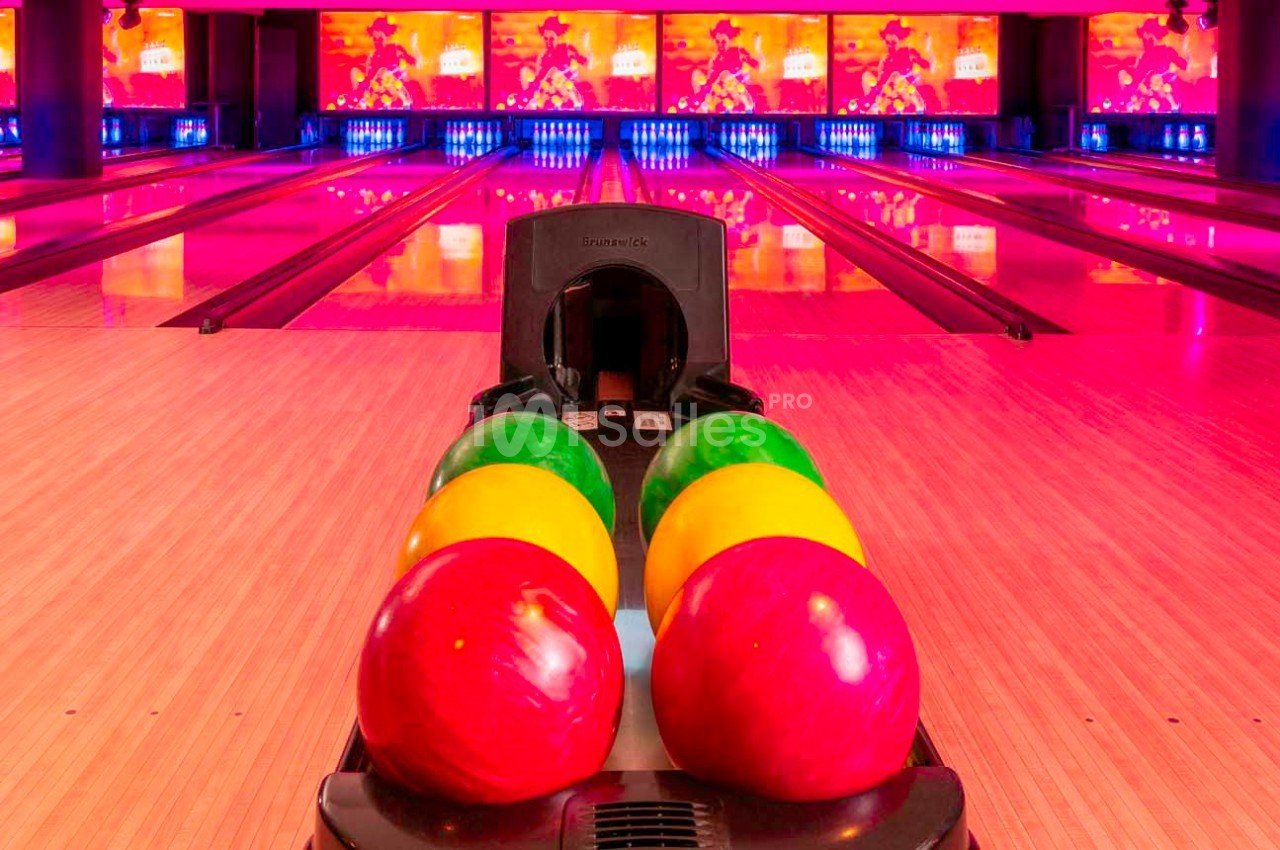 Quatre boules de bowling colorées alignées sur une rampe devant des pistes éclairées par des lumières roses.