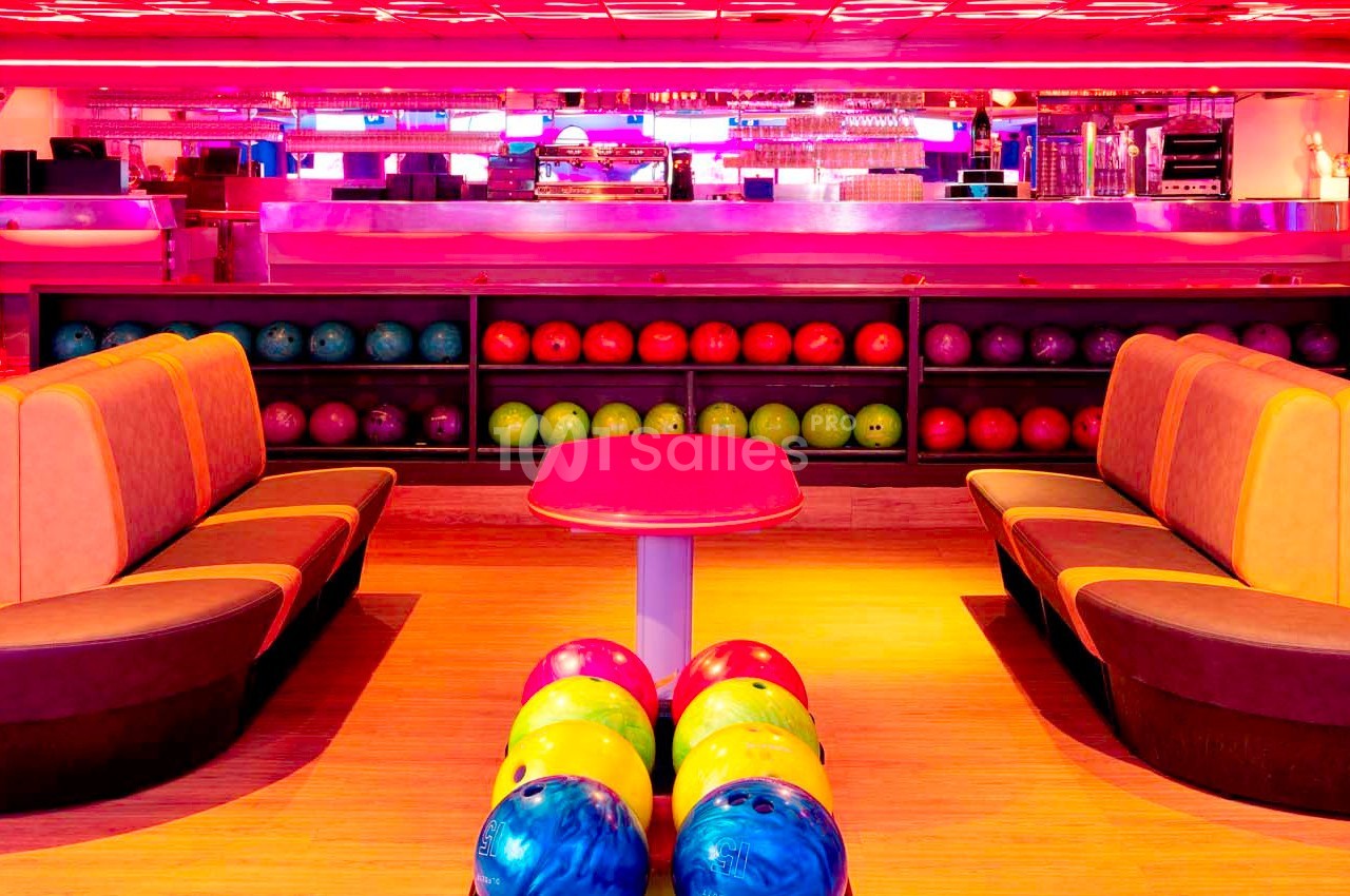 Salle de bowling colorée avec banquettes, table centrale, rangée de boules multicolores et éclairage néon.