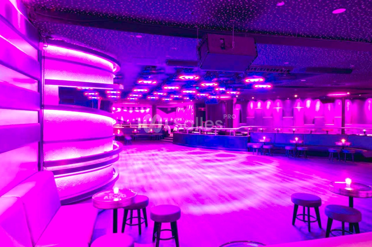 Salle de danse vide éclairée par des lumières violettes, avec des tables et des sièges disposés autour.