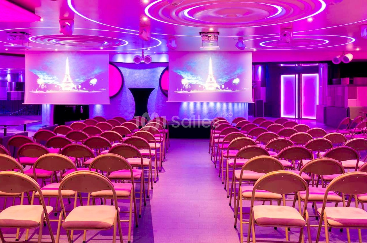 Salle de conférence avec rangées de chaises pliantes, éclairage rose et écrans de projection affichant des images.