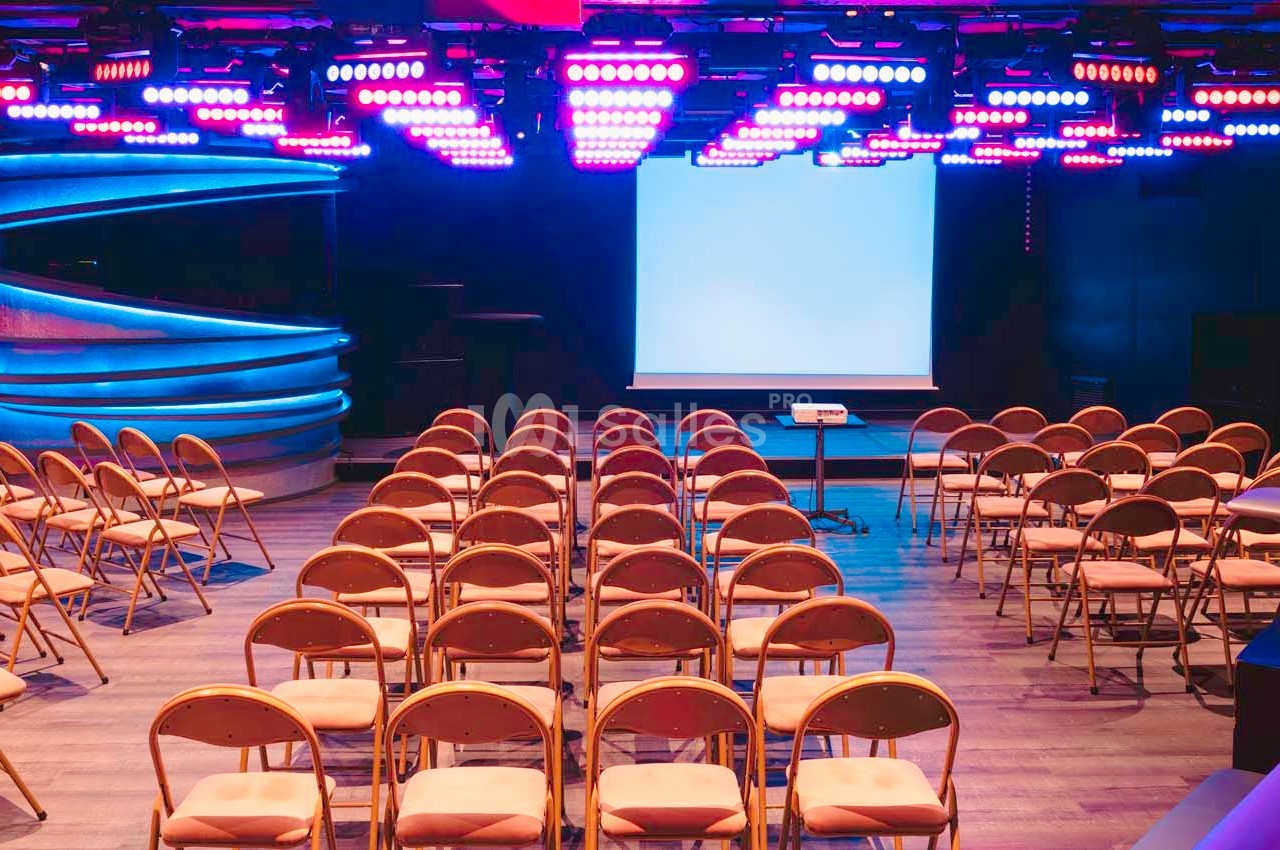 Salle de conférence avec des rangées de chaises pliantes face à un écran de projection, éclairée par des lumières colorées.