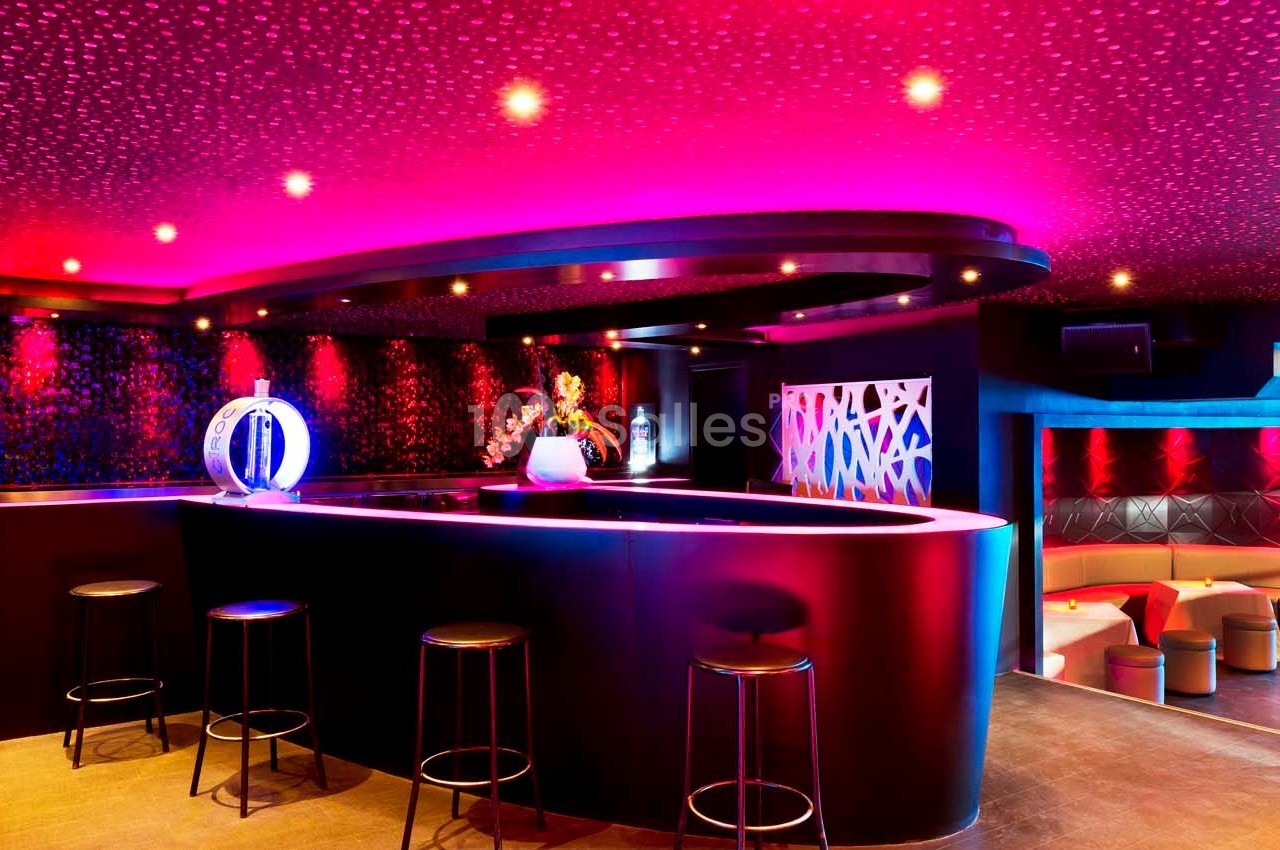Bar moderne avec éclairage coloré, tabourets noirs, comptoir incurvé et décorations lumineuses dans une ambiance tamisée.