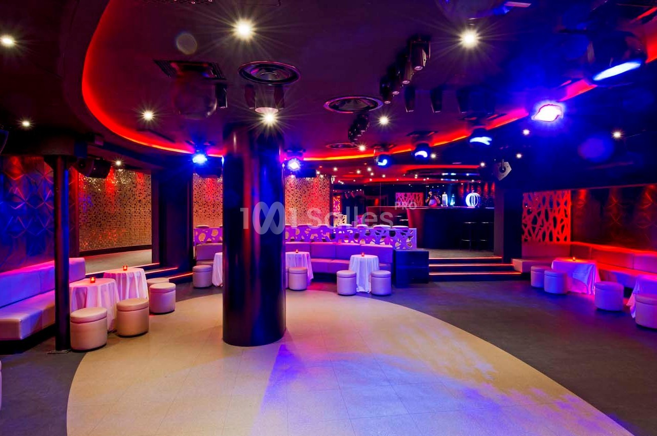 Salle de club nocturne avec éclairage rouge et violet, banquettes blanches et tables basses disposées autour.