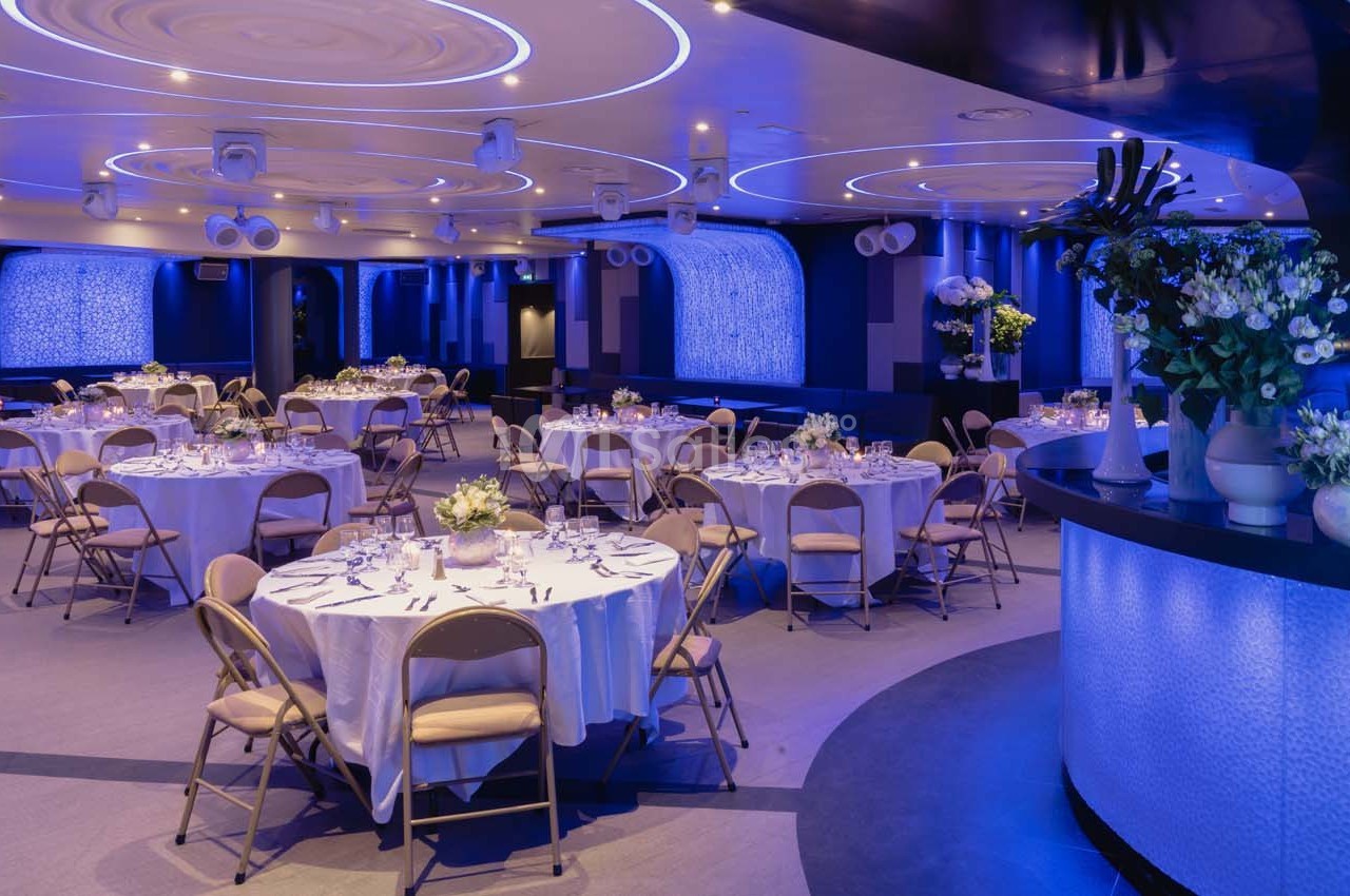 Salle de réception élégante avec tables rondes dressées, éclairage bleu et décoration florale.
