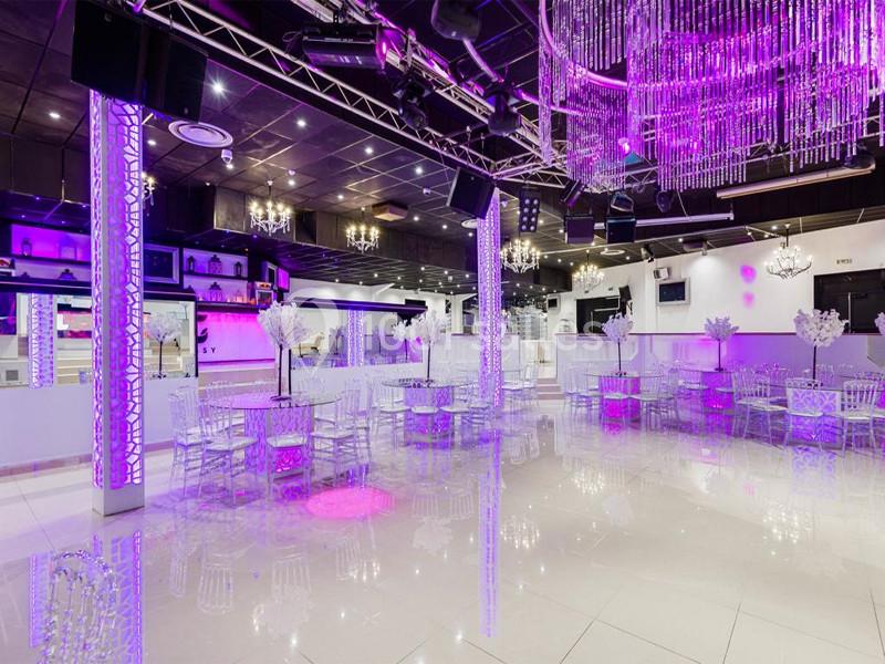 Salle de réception moderne avec tables, chaises transparentes et éclairage violet sous des lustres en cristal.