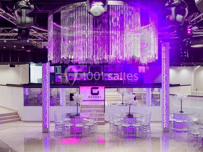 Salle de réception moderne avec tables transparentes, éclairage violet et grand lustre en cristaux suspendu.