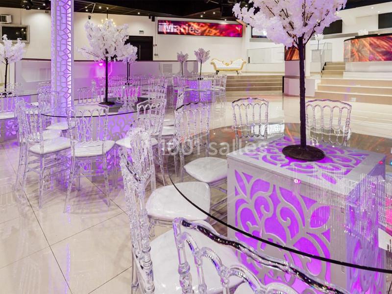 Salle de réception décorée avec des tables en verre, chaises transparentes et arbres artificiels illuminés.
