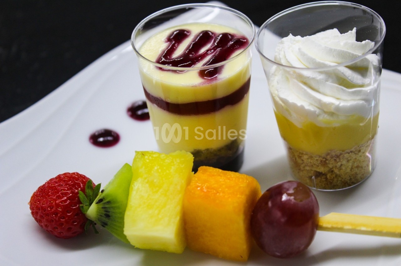 Deux verrines de dessert, l'une avec chantilly, l'autre avec coulis rouge, accompagnées d'une brochette de fruits.