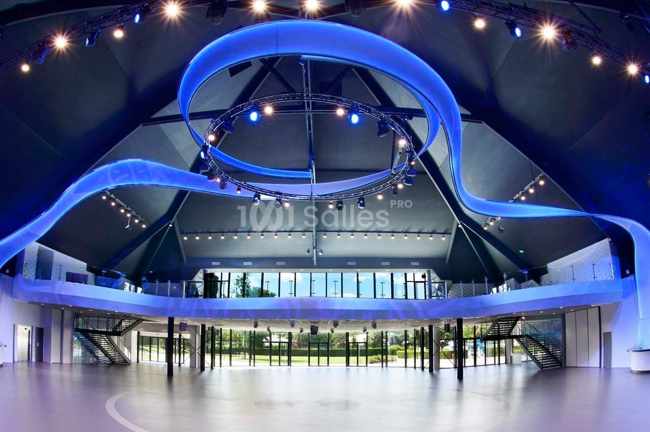Vue intérieure d'une grande salle moderne avec éclairage bleu, mezzanine vitrée et larges baies donnant sur un espace vert.