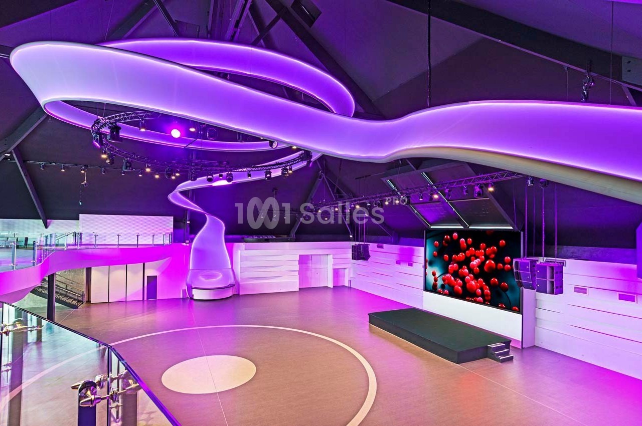 Salle moderne avec éclairage violet, écran affichant des sphères rouges et espace ouvert avec scène et balustrade.