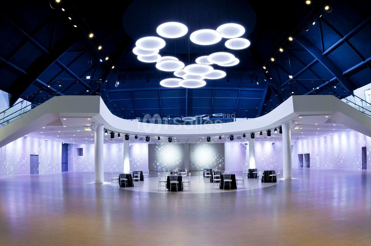 Salle spacieuse et moderne avec un éclairage suspendu circulaire, des murs blancs et un sol en bois clair.