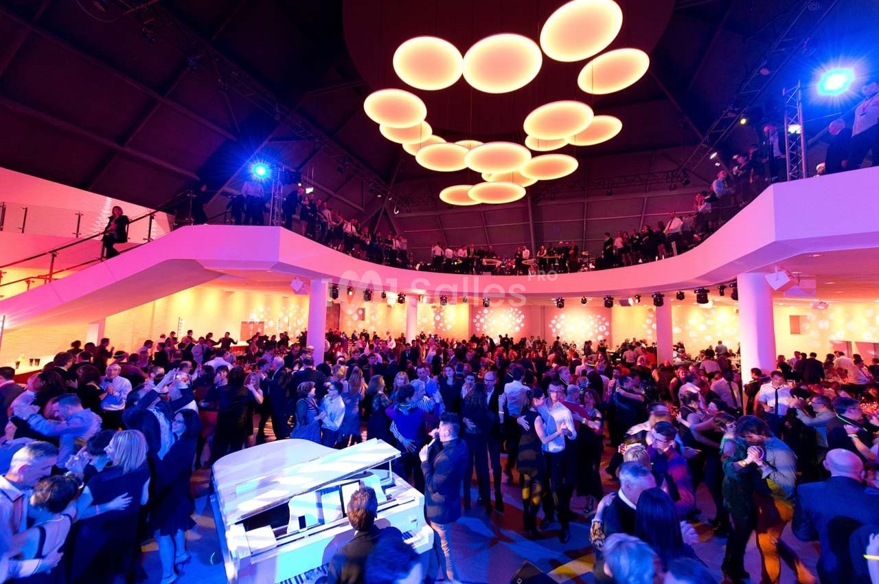 Salle de réception animée avec de nombreuses personnes dansant sous un éclairage coloré et un piano blanc au premier plan.