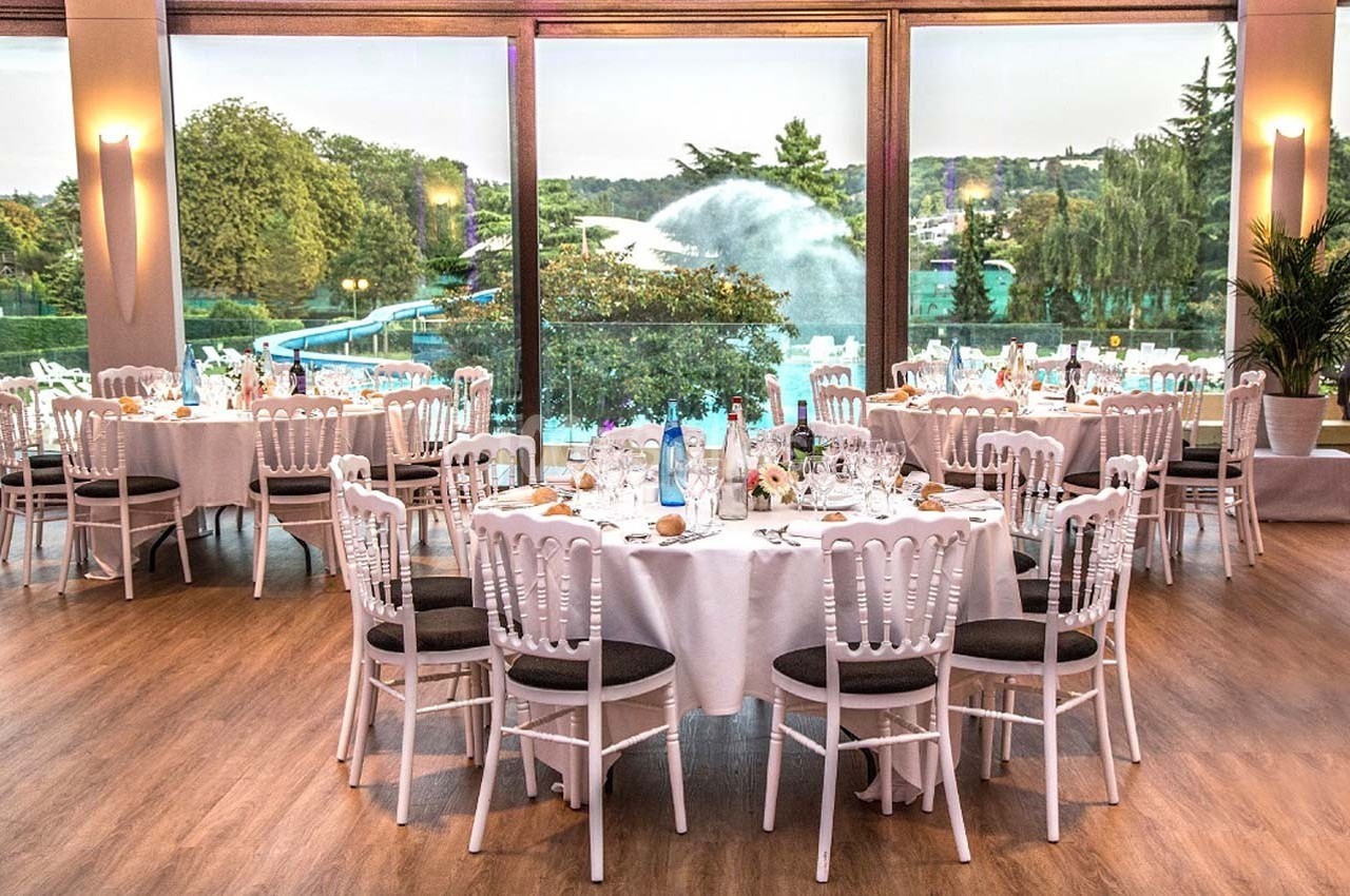 Salle de réception élégante avec tables rondes dressées, grandes baies vitrées donnant sur un jardin avec fontaine.