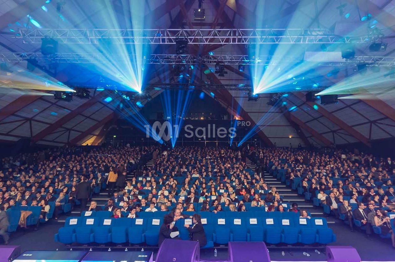 Salle comble avec un public assis, éclairée par des projecteurs bleus sous une structure en bois.