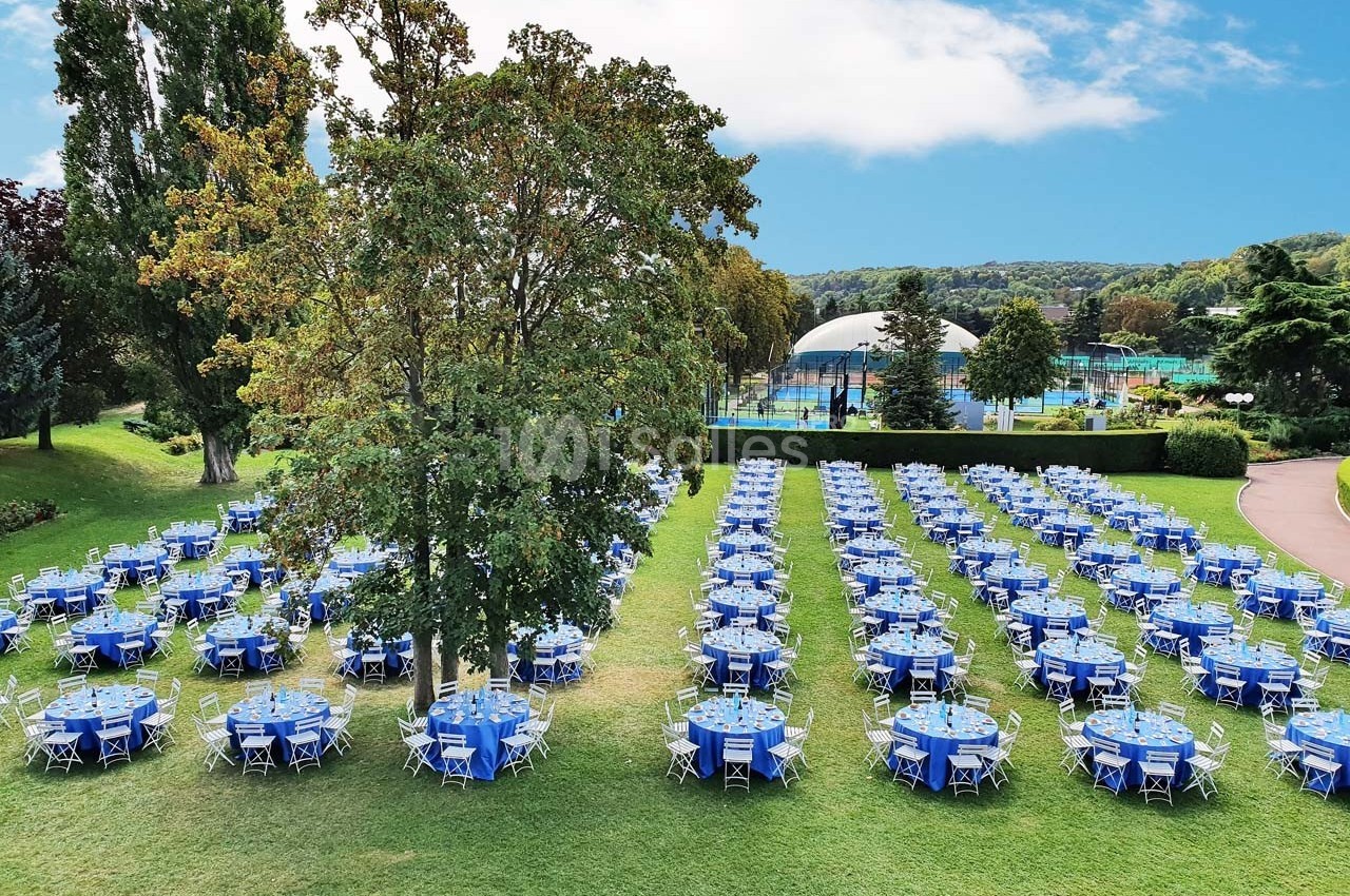 Tables rondes dressées avec nappes bleues alignées sur une pelouse, entourées d'arbres, avec un parc en arrière-plan.