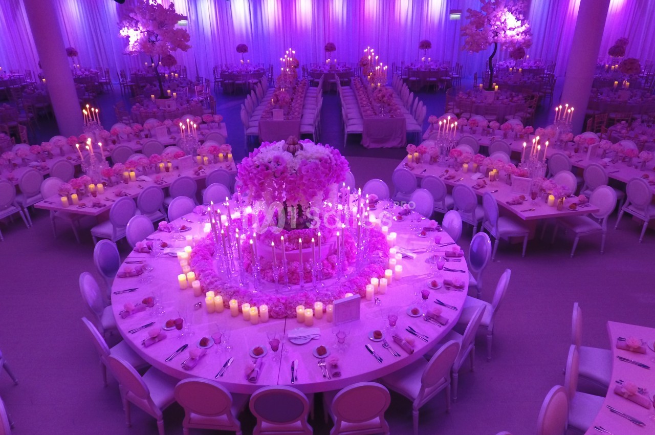 Salle de réception décorée en rose avec tables rondes, chandeliers et un grand centre de table floral illuminé.
