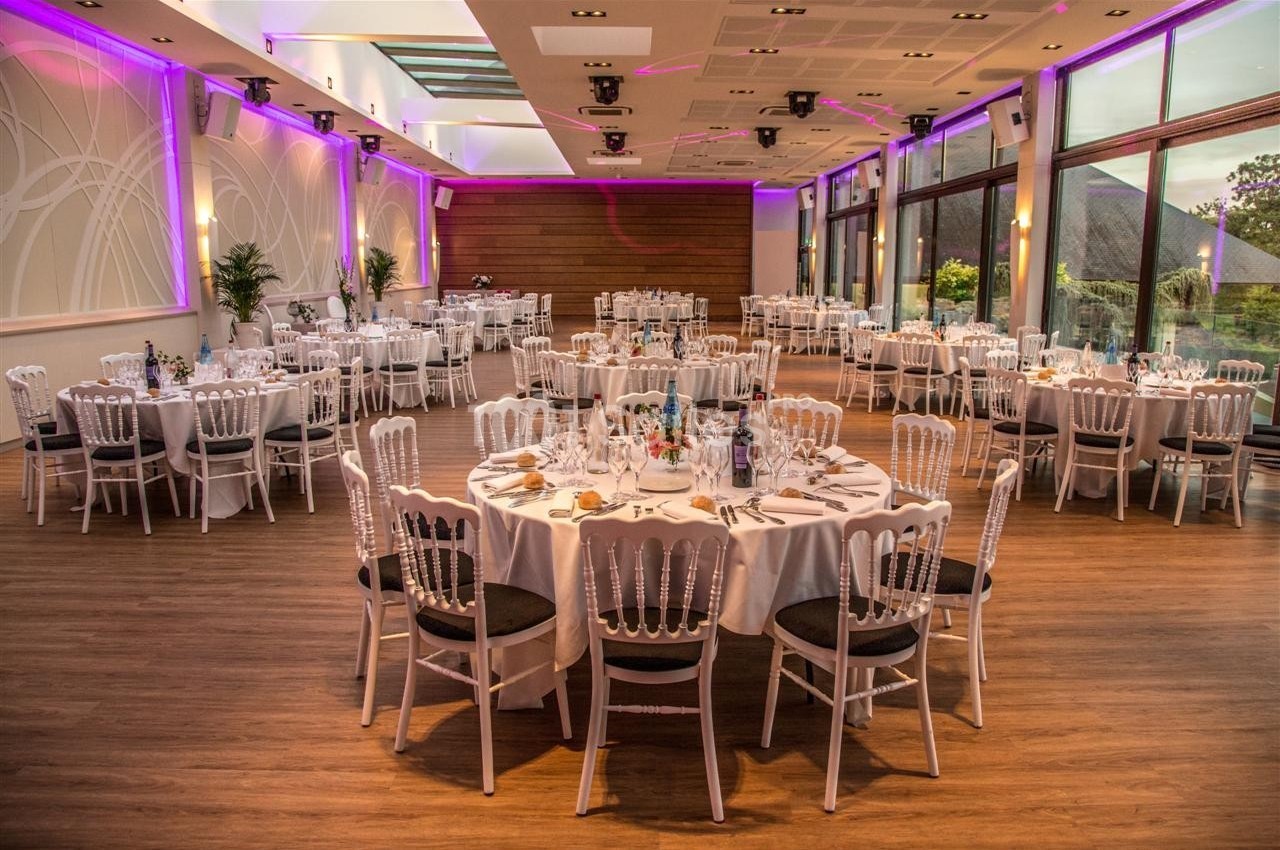 Salle de réception élégante avec tables rondes dressées, chaises blanches et éclairage tamisé.