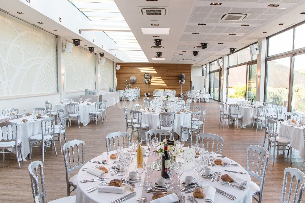 Salle de réception lumineuse avec tables rondes dressées, nappes blanches et chaises argentées.