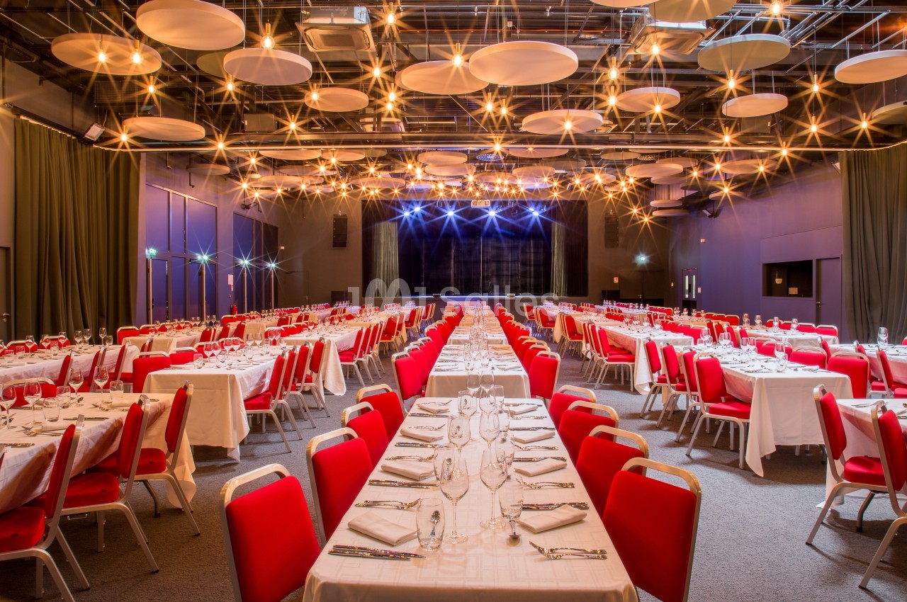 Salle de réception avec tables dressées en rangées, nappes blanches, chaises rouges et éclairage suspendu chaleureux.