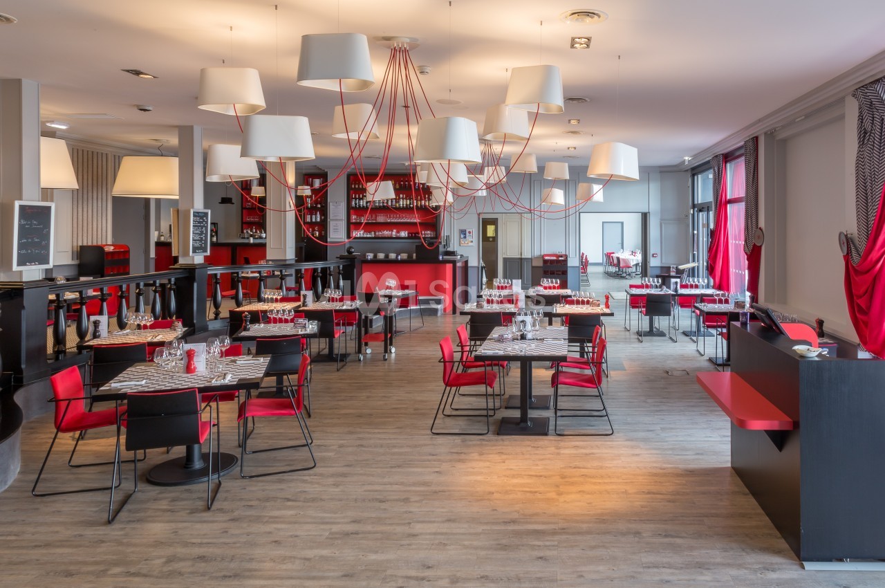 Salle de restaurant moderne avec tables dressées, chaises rouges et noires, éclairage suspendu et décor contemporain.