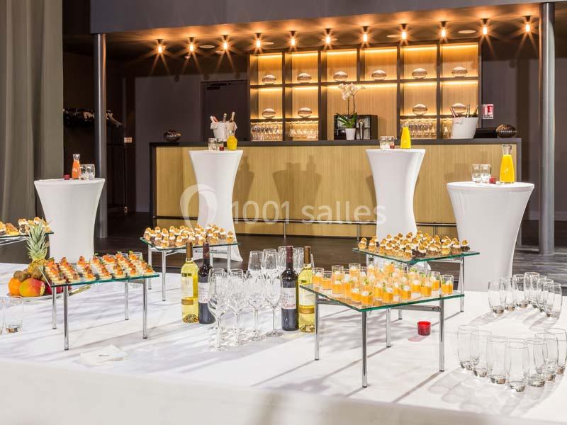 Buffet avec verrines, bouteilles de vin, jus d'orange et tables hautes dans une salle lumineuse avec un bar en arrière-plan.