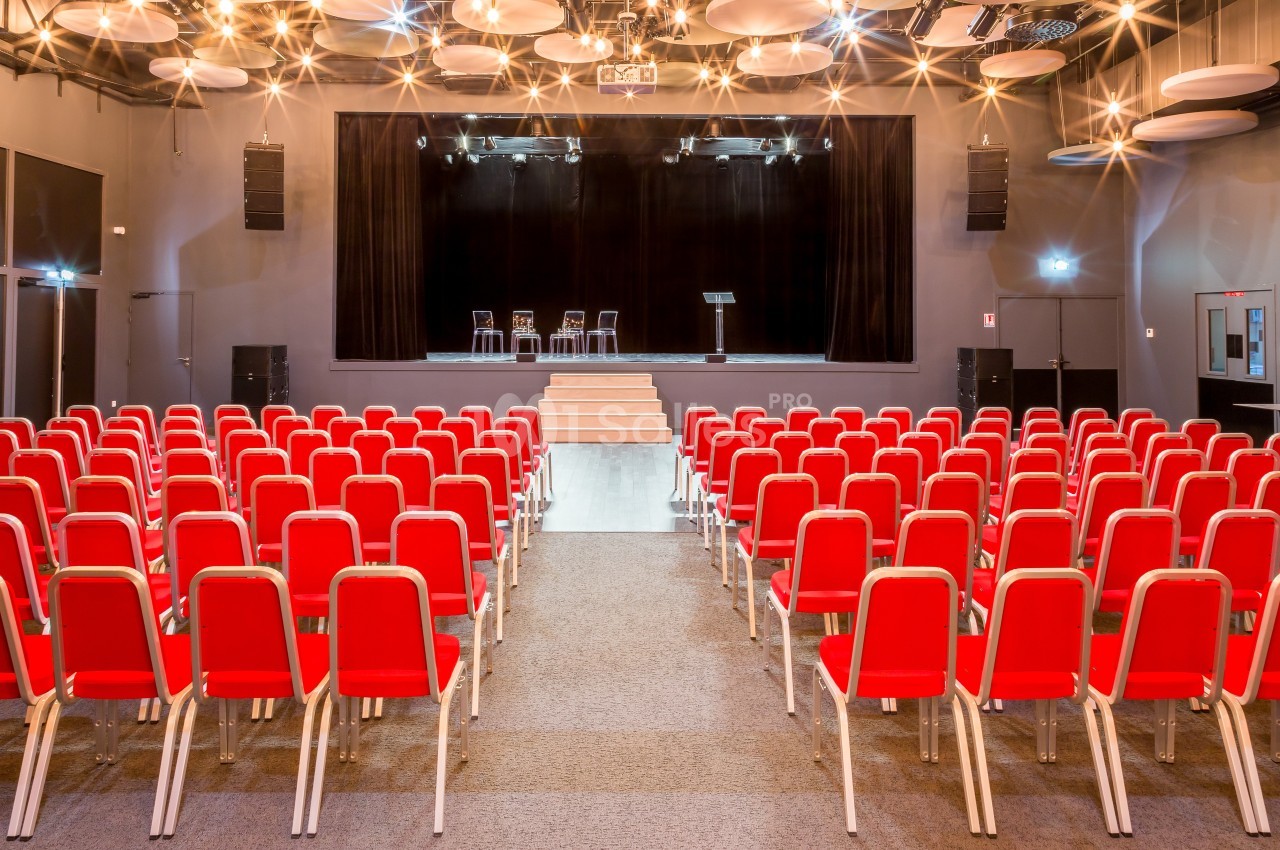 Salle de conférence avec des rangées de chaises rouges face à une scène équipée de chaises et d'un pupitre.